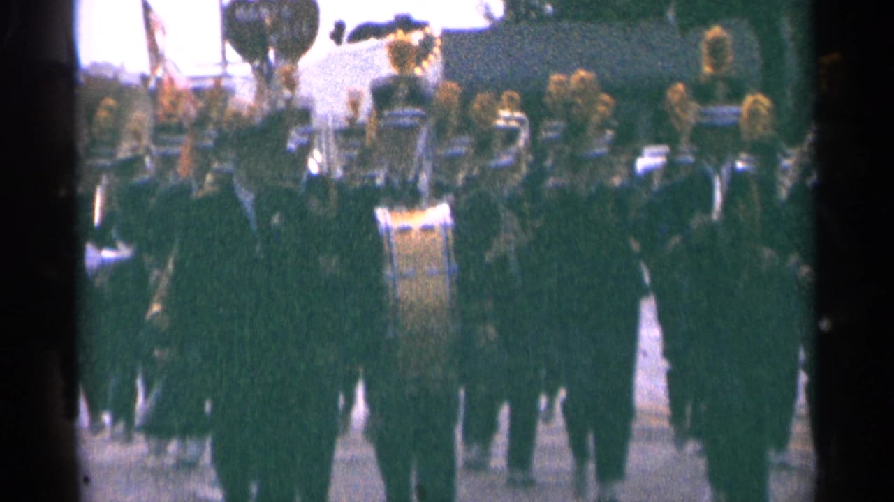 Marching band in dark uniforms advances down a street with a bass drum near the center.
