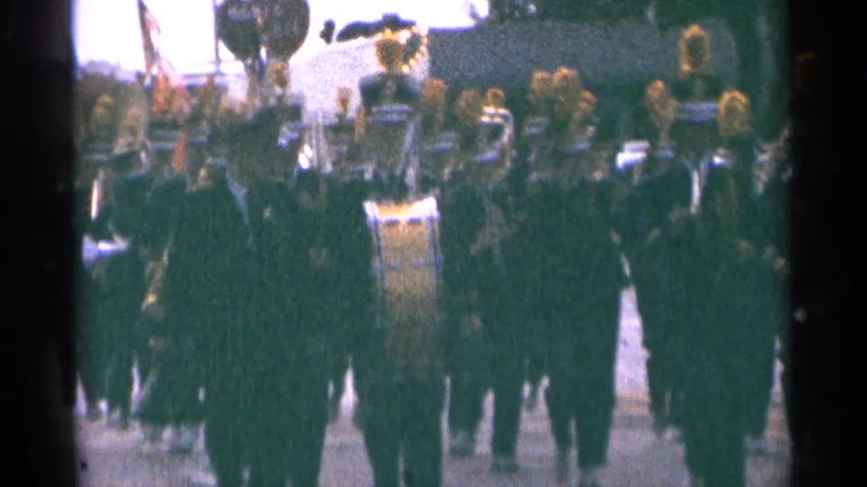 1964 Minnesota 8mm frame showing a large marching band in dark uniforms and tall gold-tasseled hats during a parade.
