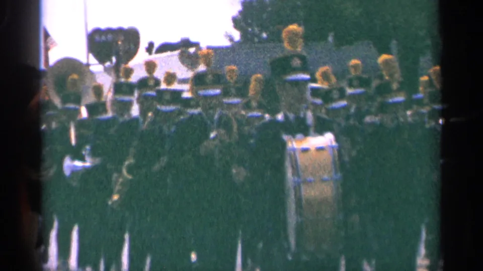 Closer 8mm view of a 1964 Minnesota parade band, with dark uniforms, tall hats, brass, and drum visible.