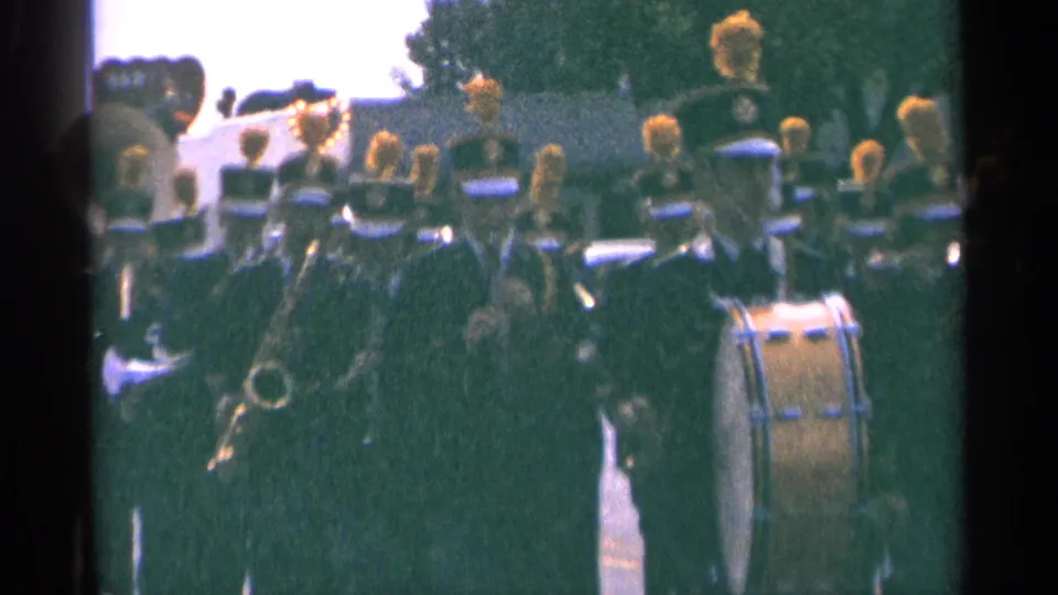 1964 Minnesota home-movie frame of a parade band moving past the camera with drum and brass instruments visible.