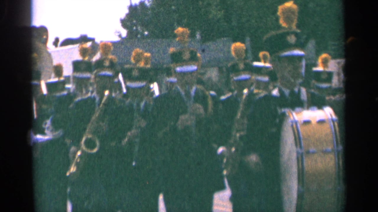 Marching band in dark uniforms approaches with yellow plumes and a bass drum visible.