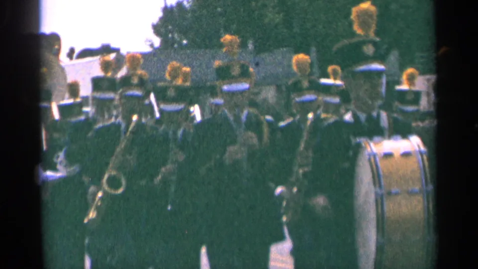 1964 Minnesota home-movie frame showing a marching band in dark uniforms with yellow plumed hats moving toward the camera.