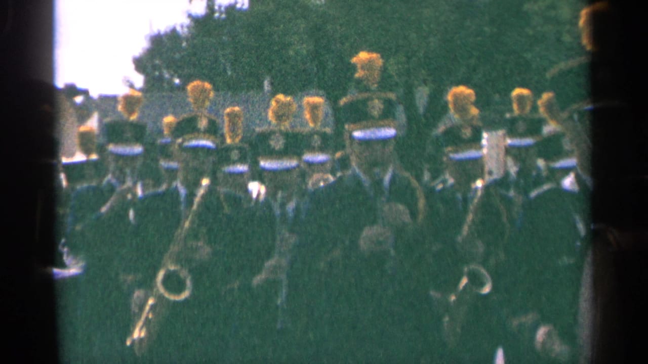 1964 Minnesota 8mm frame of uniformed band members with yellow plumes marching past trees during an outdoor event.