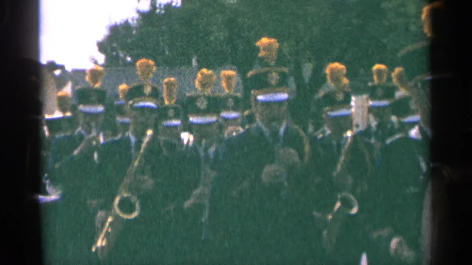 1964 Minnesota 8mm frame of uniformed band members with yellow plumes marching past trees during an outdoor event.