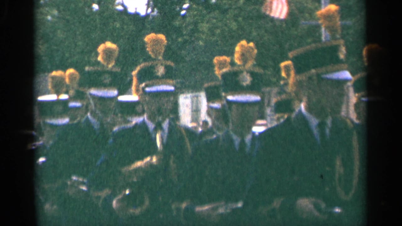 1964 Minnesota frame showing a marching band near the camera, with yellow plumes, dark uniforms, brass instruments, and a small U.S. flag.