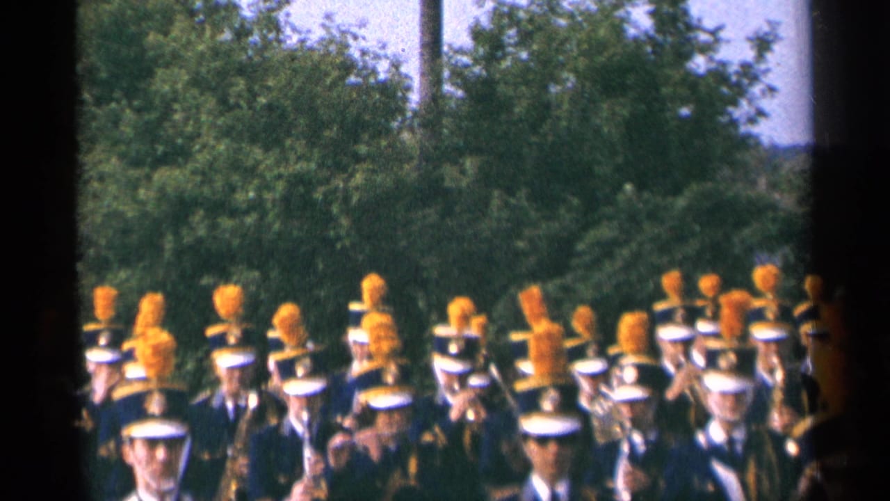Marching band members in blue uniforms and orange plumes pass trees in daylight.