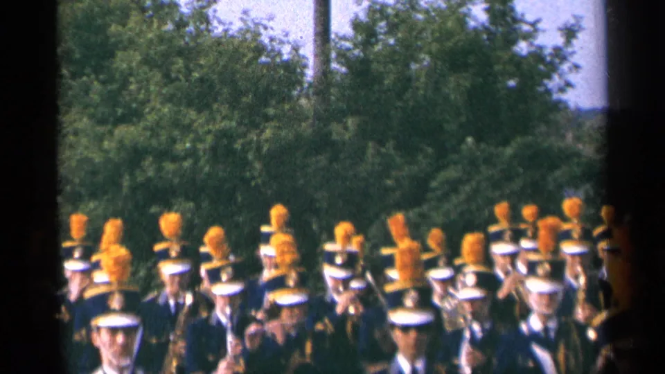 1964 Minnesota 8mm frame showing marching band members in blue uniforms, white caps, and orange plumes moving past a tree-lined roadside.
