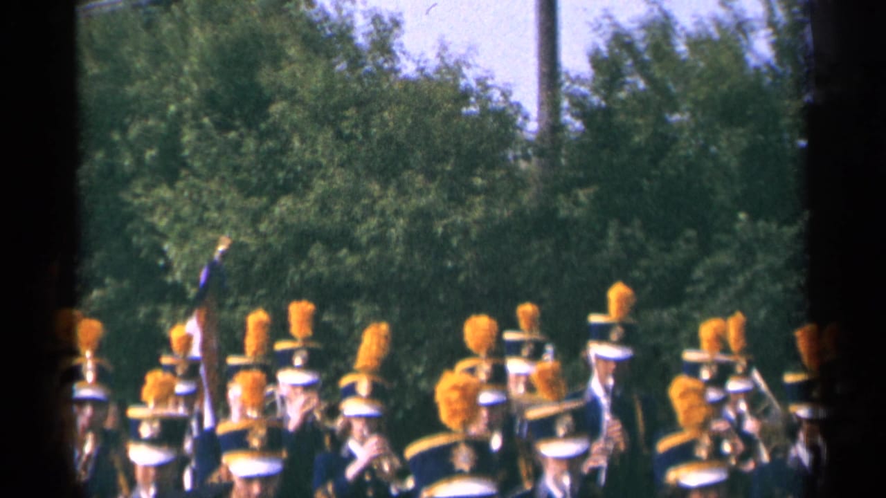1964 Minnesota parade frame with uniformed band members, orange plumes, instruments, and a flag partly visible before a leafy background.