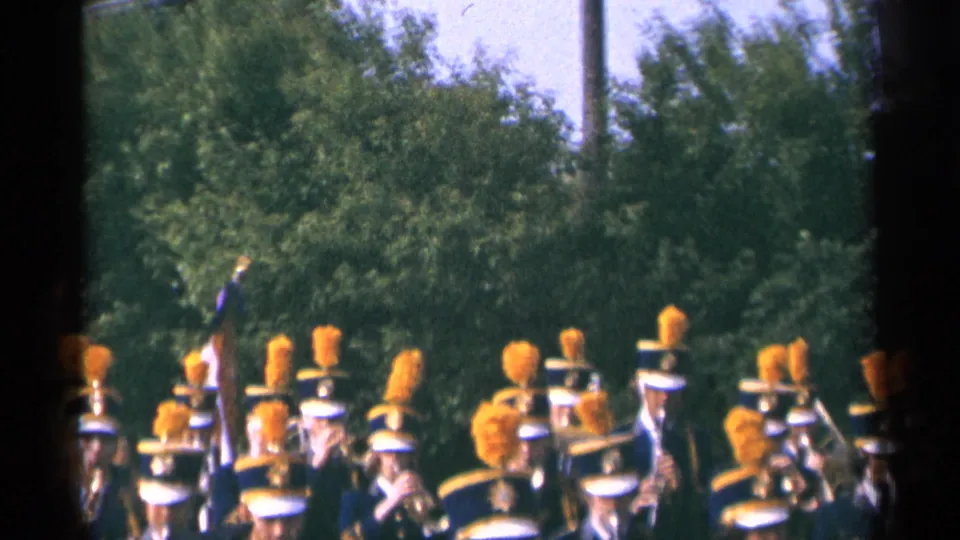 1964 Minnesota parade frame with uniformed band members, orange plumes, instruments, and a flag partly visible before a leafy background.