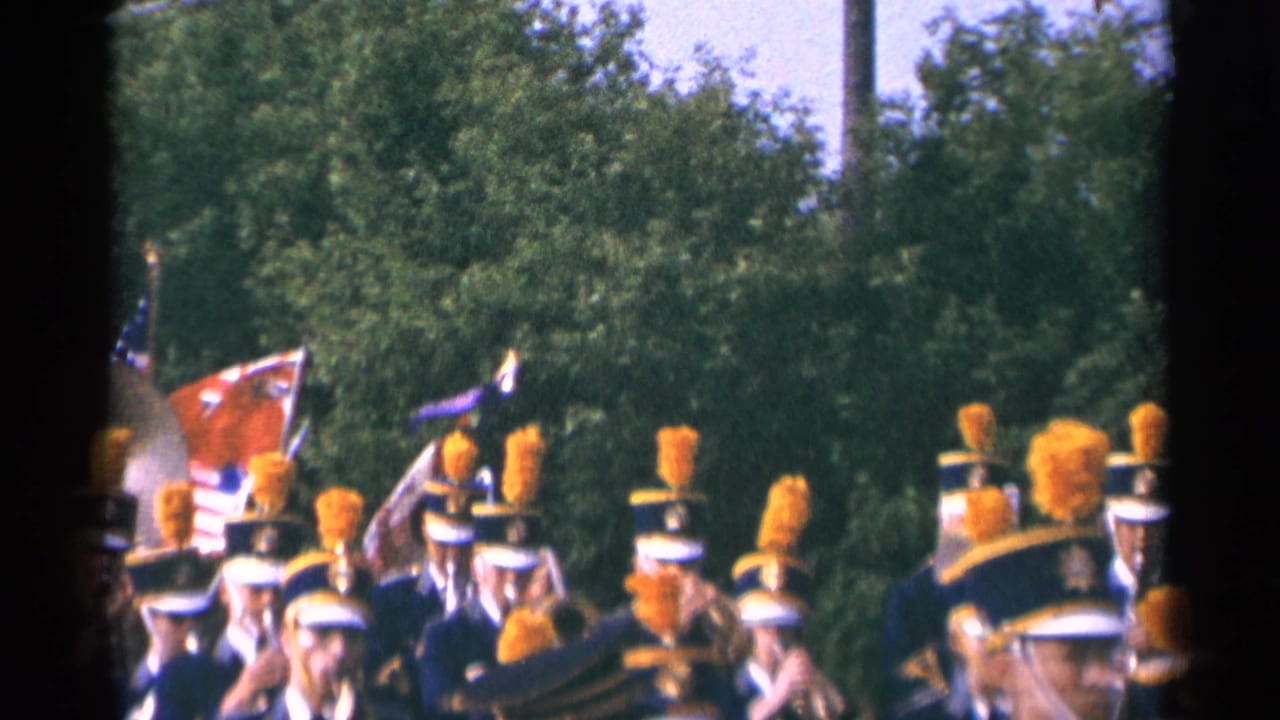 1964 Minnesota 8mm frame showing flags among marching band members in blue uniforms with orange plumes during a roadside parade.