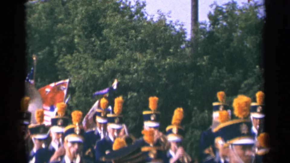 1964 Minnesota 8mm frame showing flags among marching band members in blue uniforms with orange plumes during a roadside parade.
