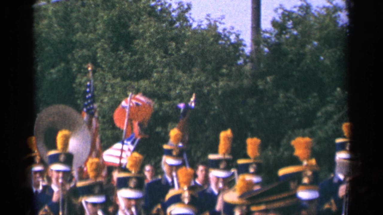 1964 Minnesota parade frame with a sousaphone, flags, and marching band members in blue uniforms with orange plumes against green trees.