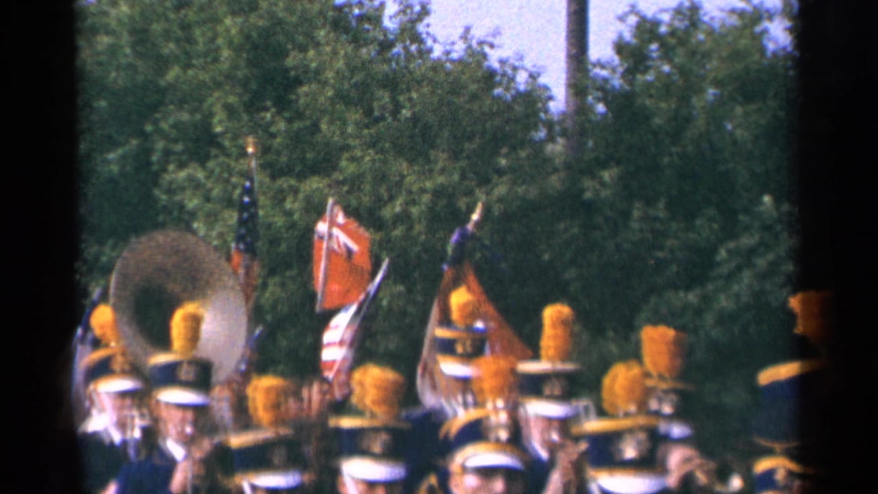 Marching band members with yellow plumes pass beneath flags on a tree-lined parade route.