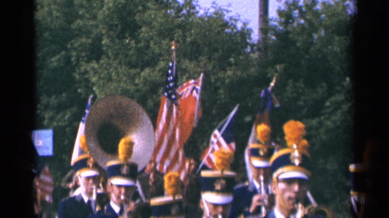 A sousaphone player and flag bearers appear among a 1964 Minnesota marching band, with spectators or marchers partly visible near the lower edge.