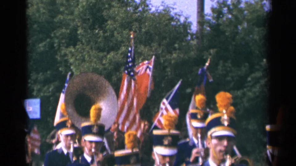 A sousaphone player and flag bearers appear among a 1964 Minnesota marching band, with spectators or marchers partly visible near the lower edge.