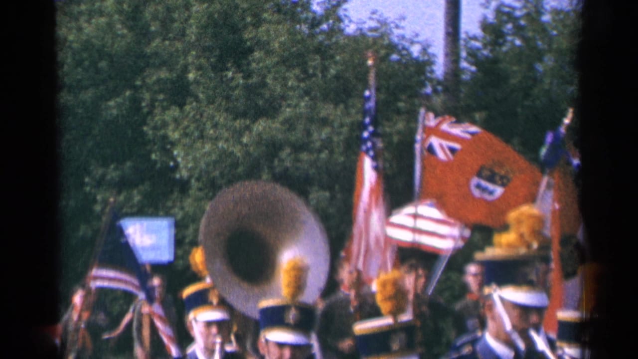 Close street-level view of the 1964 Minnesota parade band, with a sousaphone bell, yellow plumes, white hats, and a red flag crossing the frame.