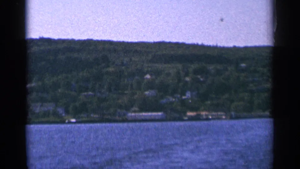 1964 Minnesota 8mm frame showing a wooded hillside and distant lakeside houses across blue water.