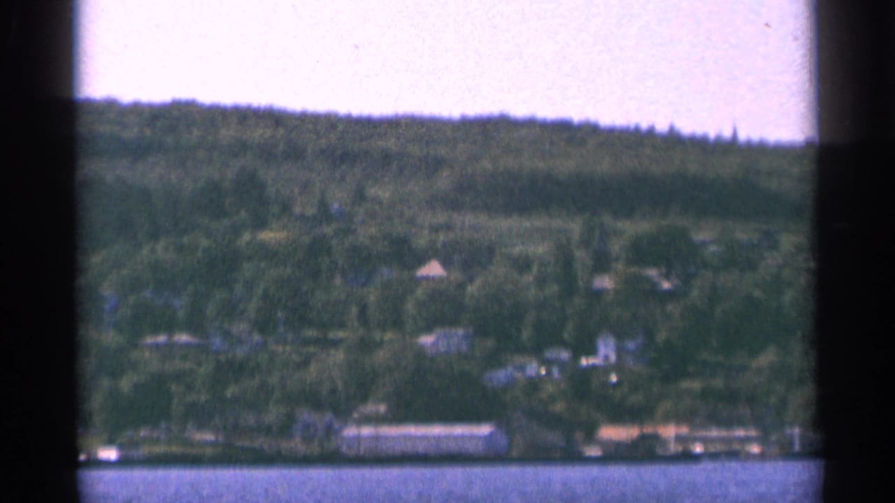1964 Minnesota home-movie frame with distant lakeside houses, trees, and low shoreline structures.