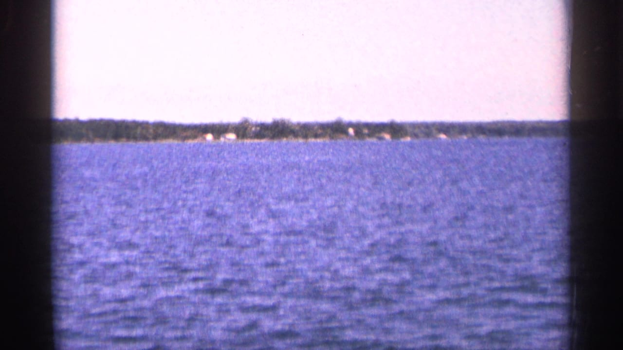 1964 Minnesota 8mm frame looking over rippled lake water toward a low wooded shoreline.