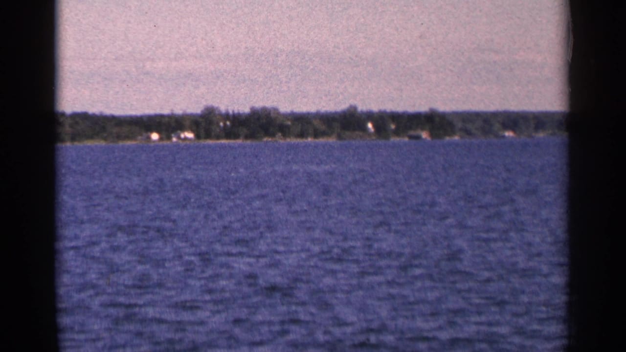 1964 Minnesota home-movie frame showing a broad lake foreground and distant wooded shore with small structures.