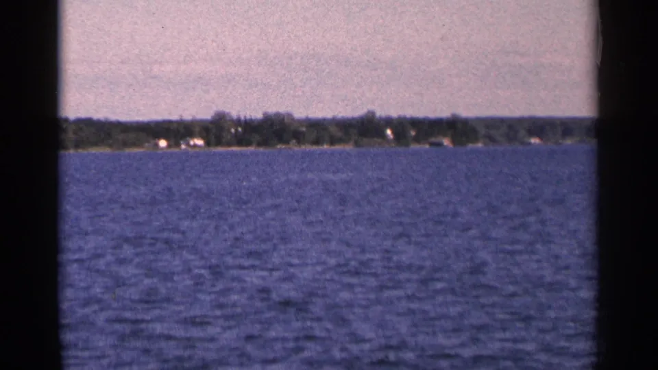 1964 Minnesota home-movie frame showing a broad lake foreground and distant wooded shore with small structures.