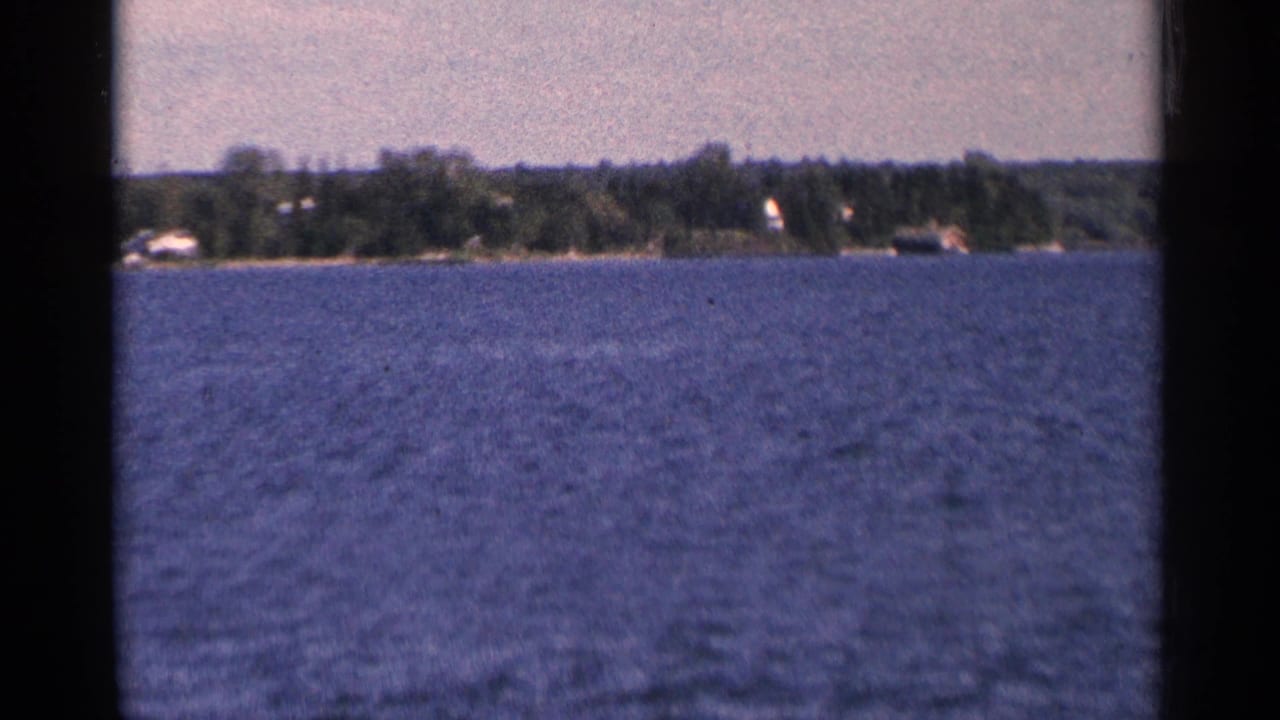 Blue rippled water with a distant wooded shoreline and small homes under a pale sky.