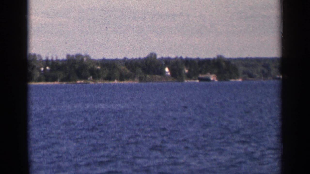 1964 Minnesota 8mm view from moving water toward a wooded shore with small distant houses and dark film edges.