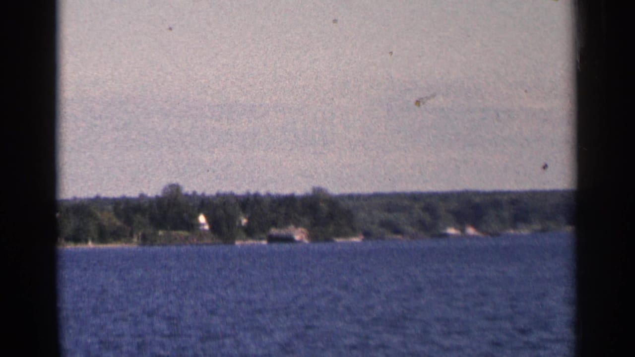 1964 Minnesota home-movie frame with rippled blue water, wooded riverbank or lakeshore, and small distant homes.