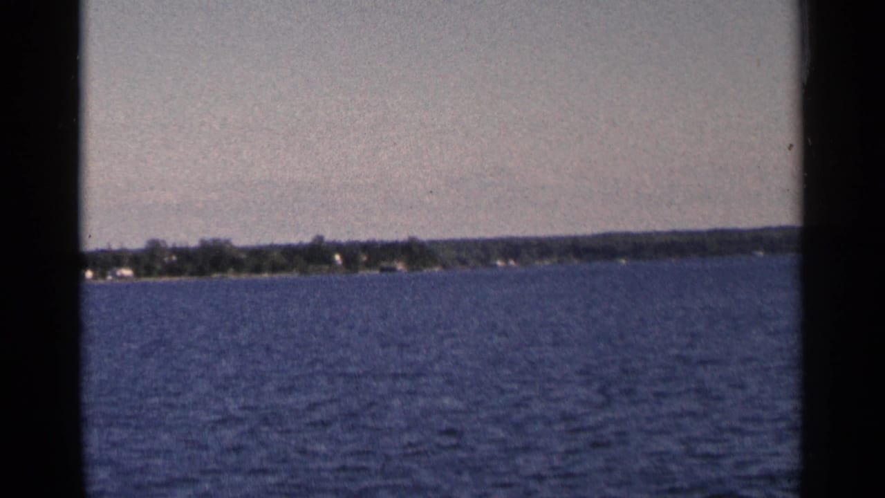 1964 Minnesota 8mm shot looking across broad blue water to a low wooded shoreline with faint houses.
