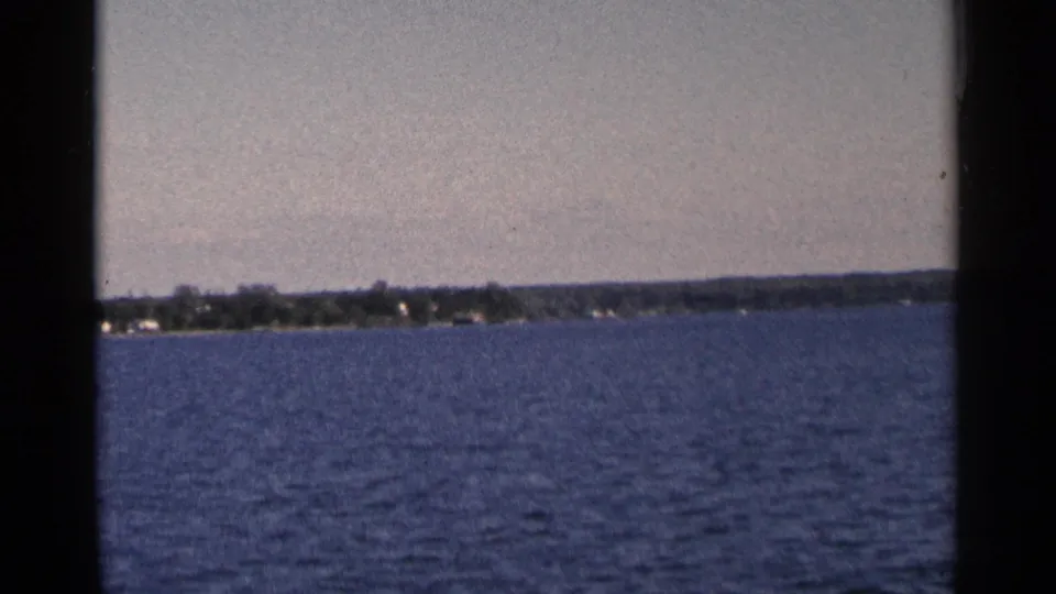 1964 Minnesota 8mm shot looking across broad blue water to a low wooded shoreline with faint houses.