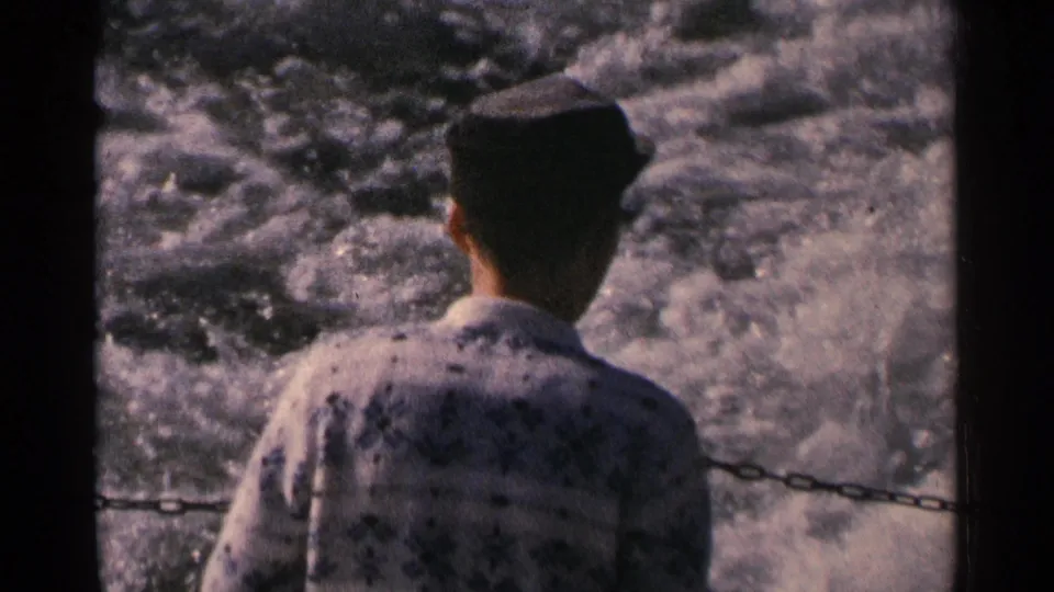A man in a dark cap and patterned shirt stands at a boat railing as the wake churns behind him in Minnesota, 1964.