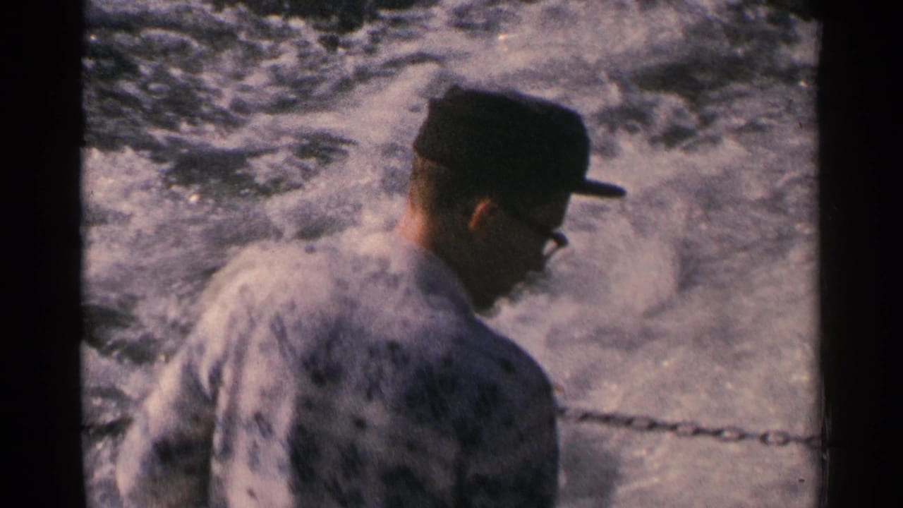 Side view of a 1964 Minnesota boat passenger wearing glasses and a dark cap beside a chain railing and foaming wake.