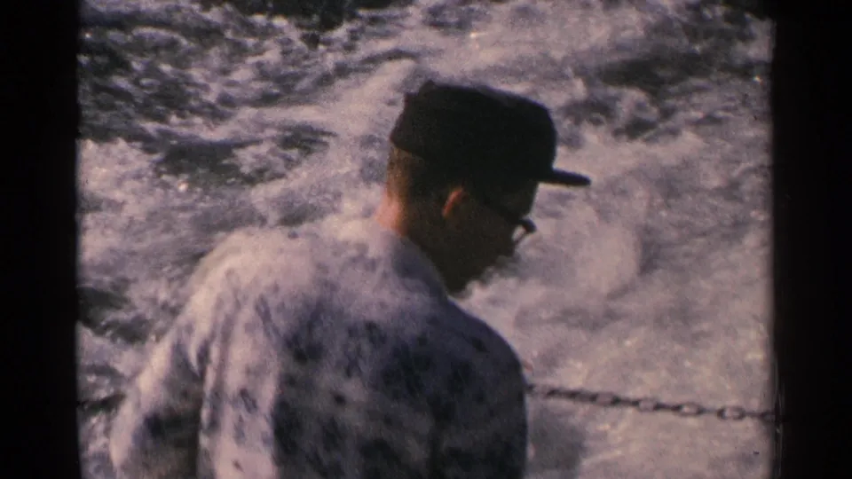 Side view of a 1964 Minnesota boat passenger wearing glasses and a dark cap beside a chain railing and foaming wake.