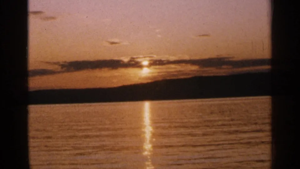 1964 Minnesota 8mm frame showing a low sun over a calm lake, with clouds, a dark shoreline, and a gold reflection on the water.