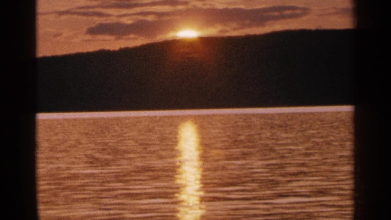 1964 Minnesota lake sunset with the sun near a dark ridge and a vertical band of light across rippled water.