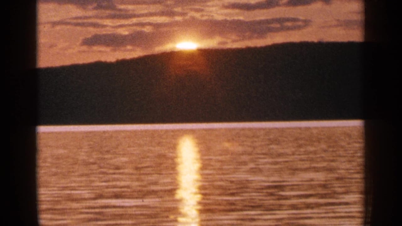 1964 Minnesota lake sunset frame with a glowing horizon, silhouetted ridge, rippled water, and warm 8mm film texture.