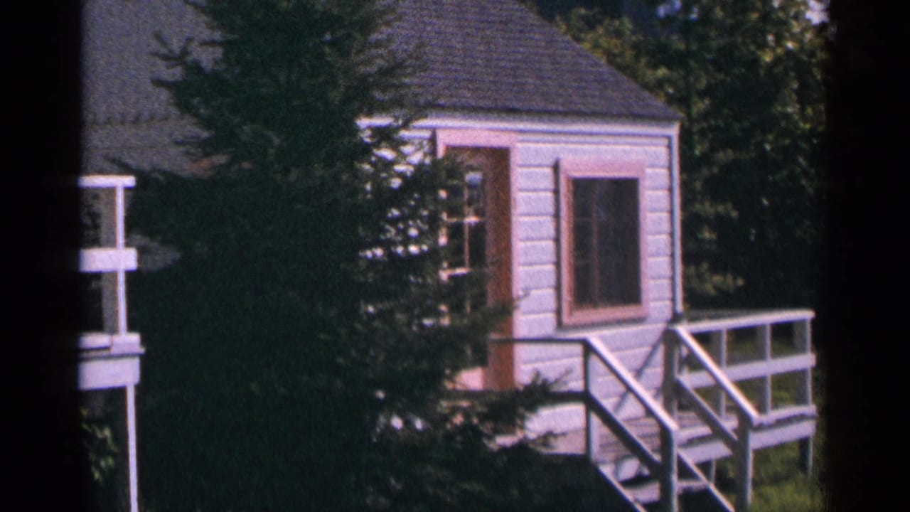 1964 Minnesota 8mm frame showing a small white cabin with pink-trimmed windows and a deck among evergreens.