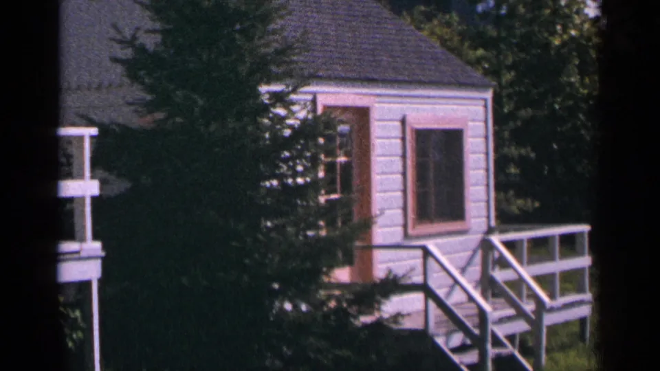 1964 Minnesota 8mm frame showing a small white cabin with pink-trimmed windows and a deck among evergreens.
