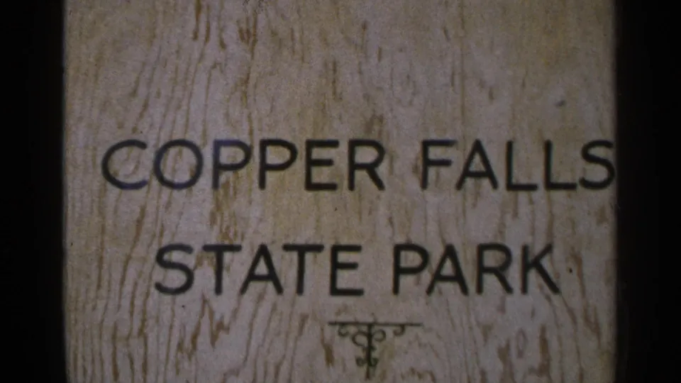 1964 home-movie frame centered on the Copper Falls State Park sign, with soft focus and faded 8mm color.