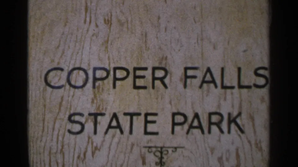 Close 8mm frame of the Copper Falls State Park sign, showing black lettering, wood texture, and film-edge darkness.