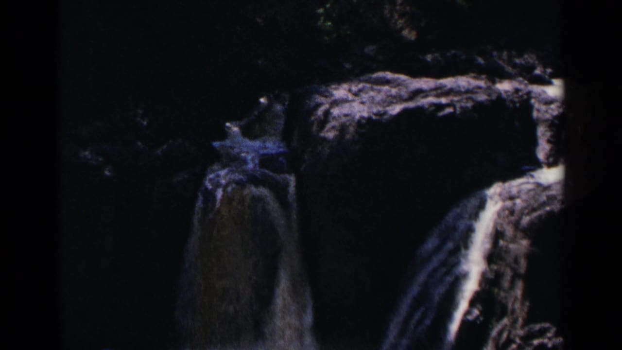 Dark 8mm frame of water falling over rocky ledges in a shaded park setting.