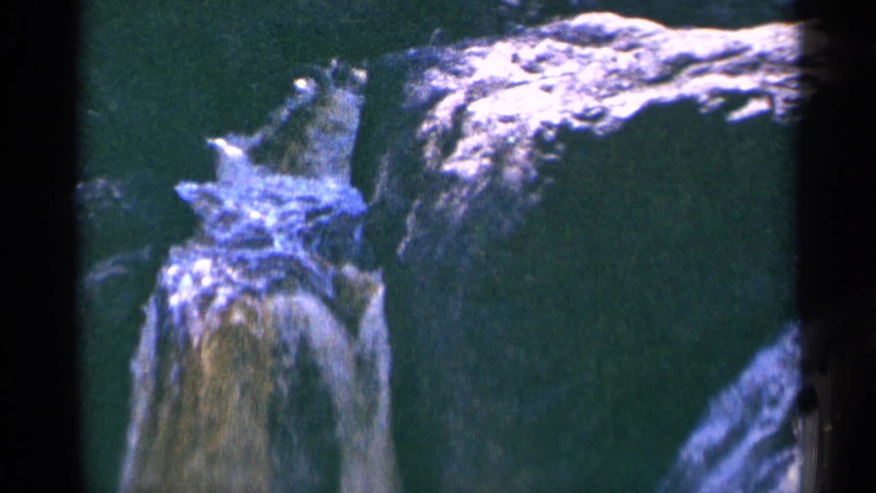 1964 Minnesota waterfall detail with bright white water cresting over a rocky shelf above a green pool, filmed with 8mm grain.