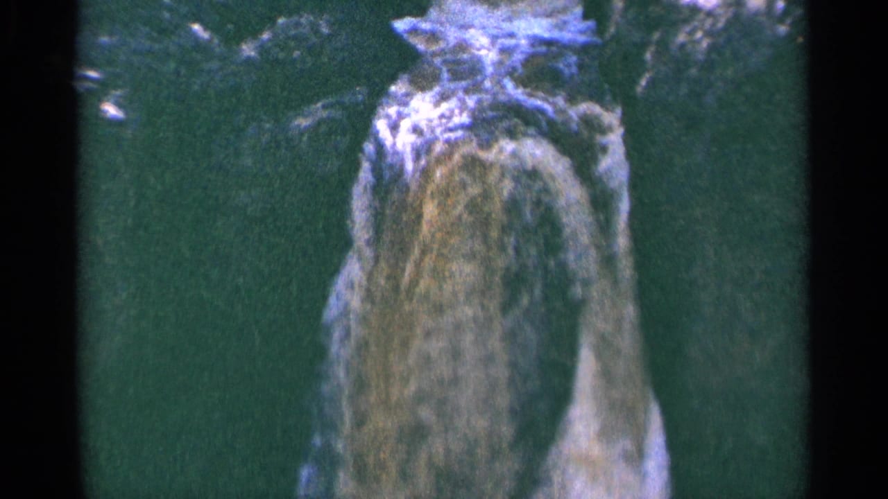 1964 Minnesota view of water cascading down a rocky face into green water, framed by dark 8mm gate edges.
