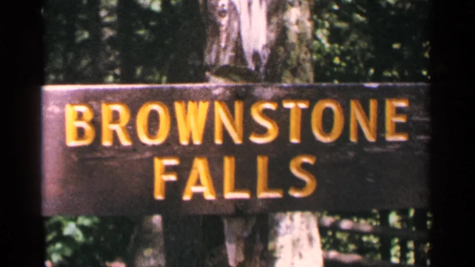 The Brownstone Falls sign remains centered in a 1964 8mm frame, with trees and foliage visible behind the board.