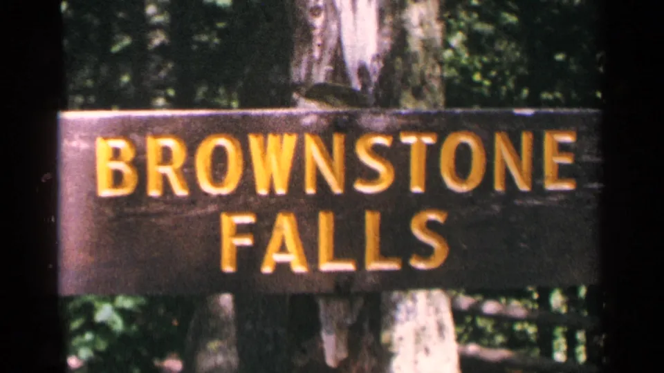 1964 Minnesota 8mm frame showing a wooden Brownstone Falls sign with yellow letters in a shaded wooded setting.