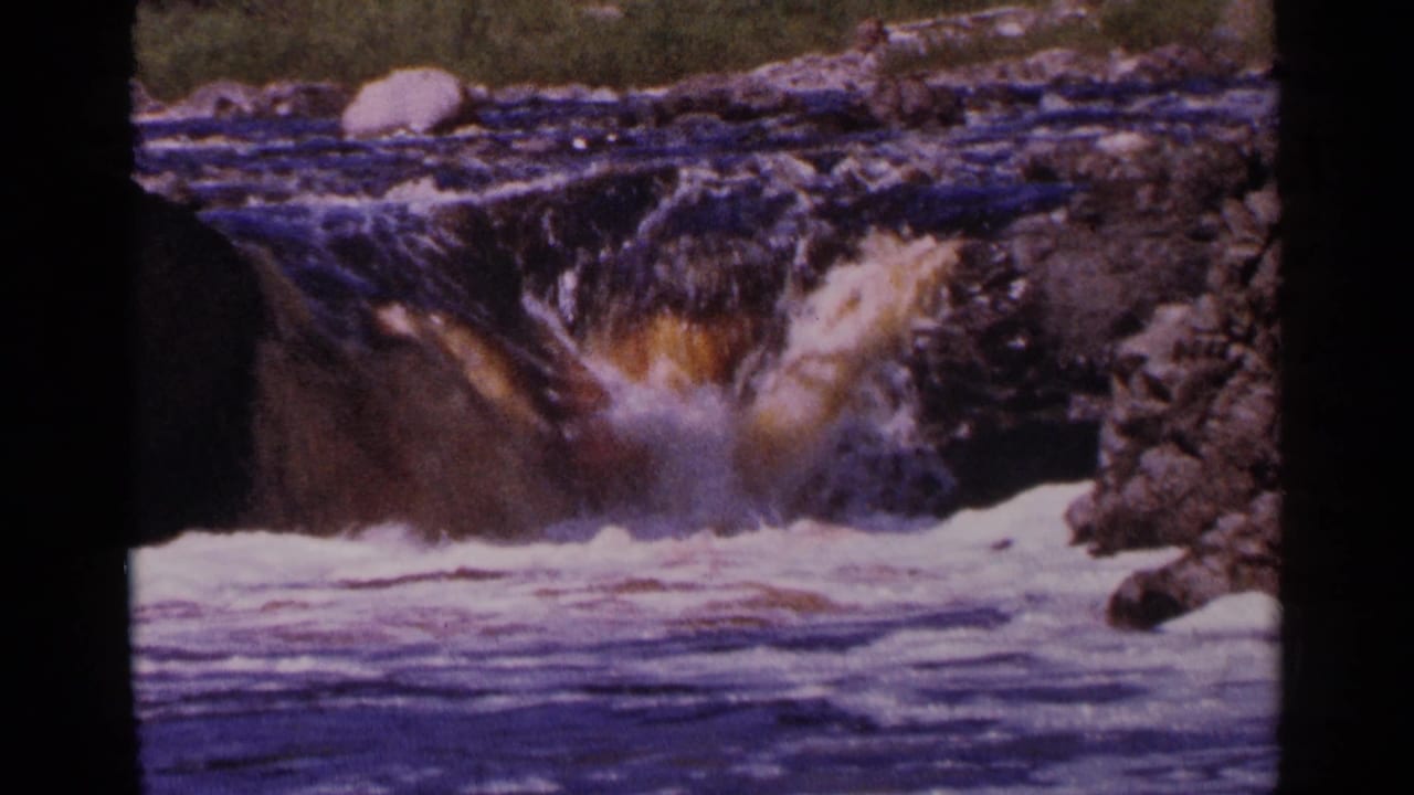 1964 Minnesota 8mm frame of a rocky river drop with foamy white water, dark current, and a pale boulder above the rapids.