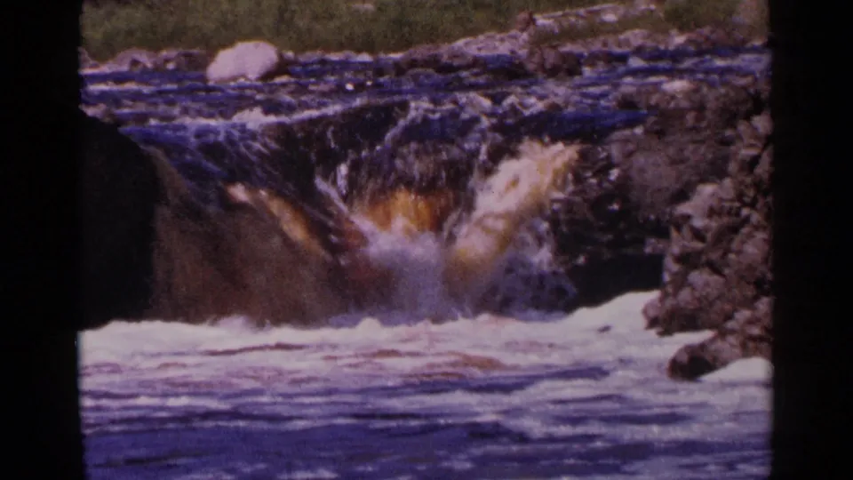 1964 Minnesota 8mm frame of a rocky river drop with foamy white water, dark current, and a pale boulder above the rapids.