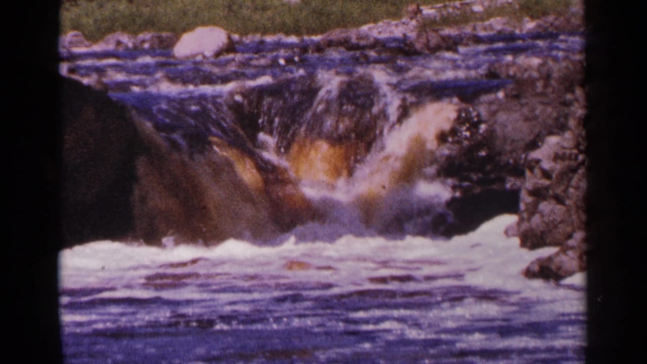 1964 Minnesota 8mm frame showing turbulent rapids pouring over rocks, with brown-purple film color shift and frothy water below.