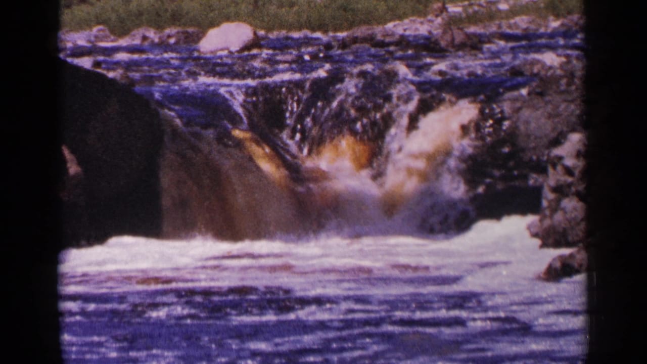 1964 Minnesota 8mm frame of white water rushing over a rocky drop, with dark current in the foreground and a boulder near the upper bank.
