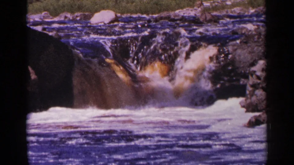 1964 Minnesota 8mm frame of white water rushing over a rocky drop, with dark current in the foreground and a boulder near the upper bank.